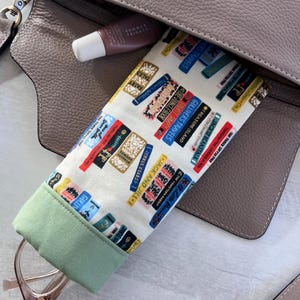 May include: A green and white fabric eyeglasses case with a book print pattern. The case is lined with a green fabric and has a zipper closure. The case is in a brown leather handbag.