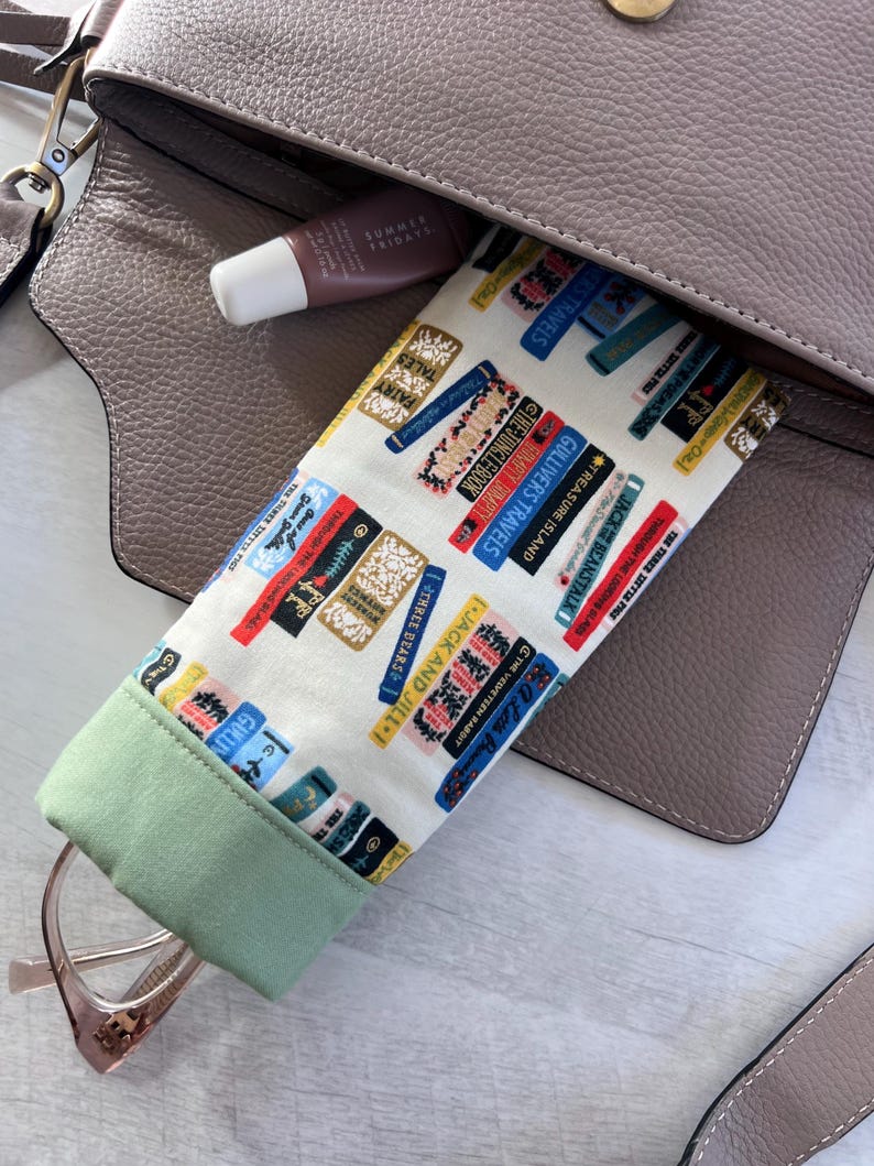 May include: A green and white fabric eyeglasses case with a book print pattern. The case is lined with a green fabric and has a zipper closure. The case is in a brown leather handbag.