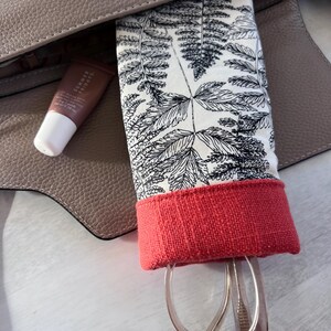 May include: A red and white patterned glasses case with a fern design. The case is in a brown leather bag and has a pair of glasses inside.