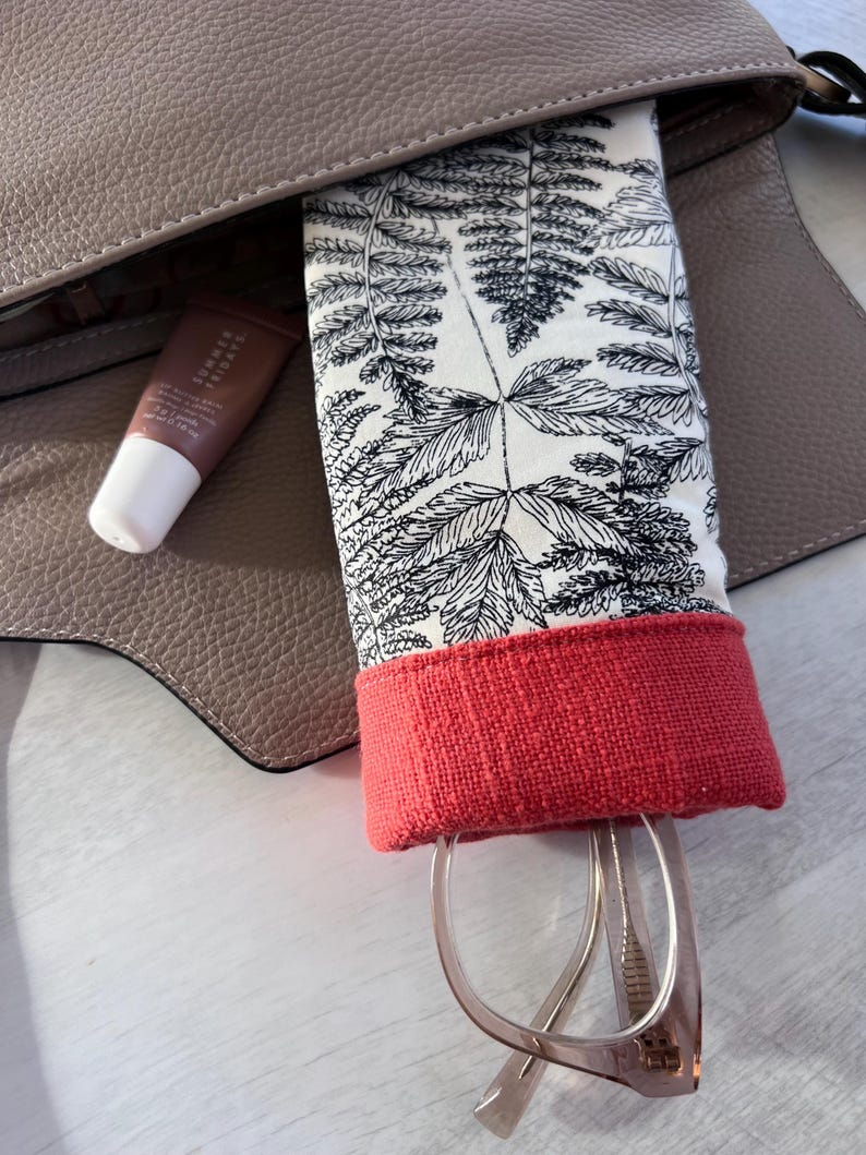 May include: A red and white patterned glasses case with a fern design. The case is in a brown leather bag and has a pair of glasses inside.