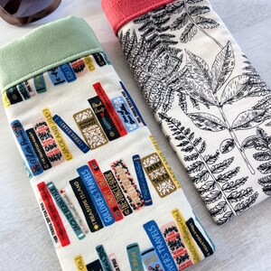 May include: Two fabric eyeglass cases. One case has a white background with a black fern pattern and a coral colored top. The other case has a white background with a pattern of colorful books and a light green top.