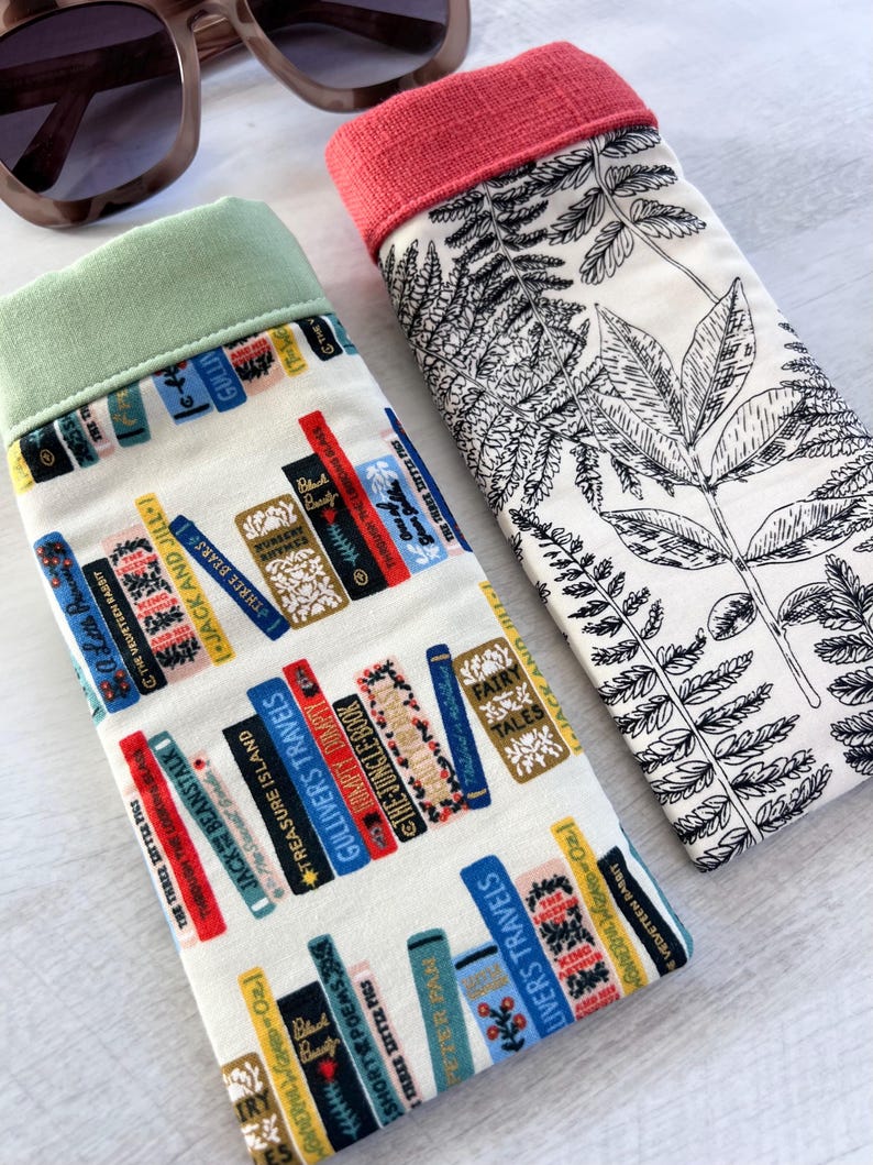 May include: Two fabric eyeglass cases. One case has a white background with a black fern pattern and a coral colored top. The other case has a white background with a pattern of colorful books and a light green top.