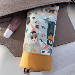 May include: A light blue and yellow fabric pouch with a colorful print of people and palm trees. The pouch is partially inside a brown leather purse.