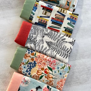 May include: Five fabric swatches with different patterns. The top swatch is a light green fabric with a white and brown pattern of people relaxing on a beach. The second swatch is a white fabric with a black and blue pattern of stacked books. The third swatch is a white fabric with a black pattern of ferns. The fourth swatch is a white fabric with a colorful pattern of flowers and tulips. The bottom swatch is a light blue fabric with a white pattern of circles containing flowers and butterflies.