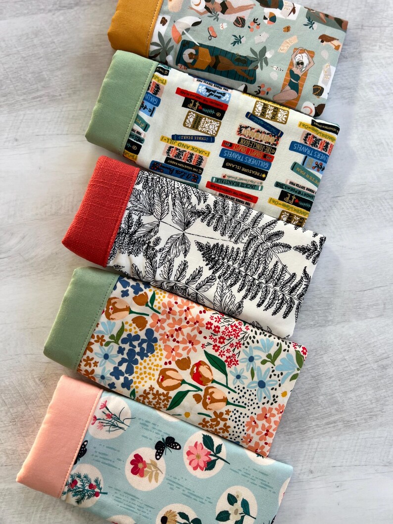 May include: Five fabric swatches with different patterns. The top swatch is a light green fabric with a white and brown pattern of people relaxing on a beach. The second swatch is a white fabric with a black and blue pattern of stacked books. The third swatch is a white fabric with a black pattern of ferns. The fourth swatch is a white fabric with a colorful pattern of flowers and tulips. The bottom swatch is a light blue fabric with a white pattern of circles containing flowers and butterflies.