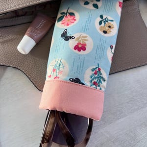 May include: A light blue and pink fabric sunglasses case with a floral print. The case is open and a pair of brown sunglasses are inside. The sunglasses have a gold metal frame and the text "DIFF" is visible on the side.