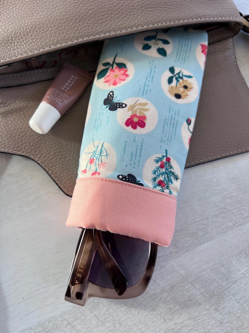 May include: A light blue and pink fabric sunglasses case with a floral print. The case is open and a pair of brown sunglasses are inside. The sunglasses have a gold metal frame and the text "DIFF" is visible on the side.