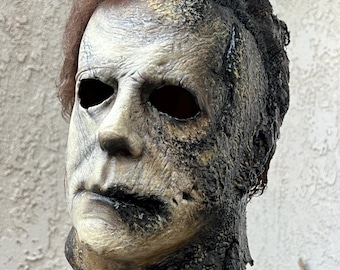 May include: A detailed Halloween mask of Michael Myers from the movie Halloween. The mask is made of latex and has a realistic, weathered look. The mask is painted in shades of brown, black, and white, and has a realistic texture. The mask is on a stand.