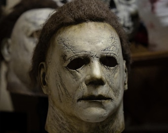 Könnte beinhalten: Eine verwitterte, cremefarbene Halloween-Maske mit dunklen Augenhöhlen und einem ernsten Gesichtsausdruck. Die Maske hat braunes Haar und sichtbare Falten, die auf Alter und Abnutzung hindeuten. Weitere ähnliche Masken sind im Hintergrund zu sehen.
