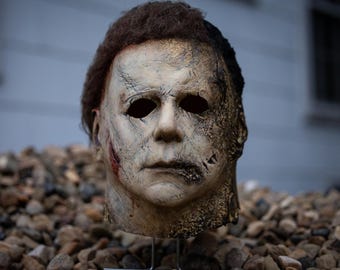 Könnte beinhalten: Eine verwitterte Michael Myers Maske mit einem blassen Gesicht, dunklen Augenhöhlen und braunem Haar. Die Maske weist sichtbare Narben und ein abgenutztes Aussehen auf, was auf ein Horrorthema hindeutet. Die Maske wird auf einem Ständer vor einem Hintergrund aus kleinen Steinen präsentiert.