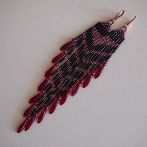 May include: A pair of long, dangling earrings featuring black and red beads. The earrings are crafted with small, round beads and larger, teardrop-shaped beads.