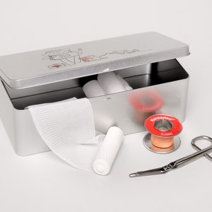 May include: A silver metal first aid kit with a cartoon illustration on the lid. The kit is open and contains bandages, gauze, a roll of adhesive tape, and a pair of scissors.