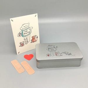 Peut inclure: Un ensemble cadeau sur le thème du rétablissement, comprenant une boîte métallique argentée avec une illustration de dessin animé, une carte de vœux avec un design similaire, deux pansements adhésifs et un cœur rouge. La carte porte l'inscription "Gute Besserung".