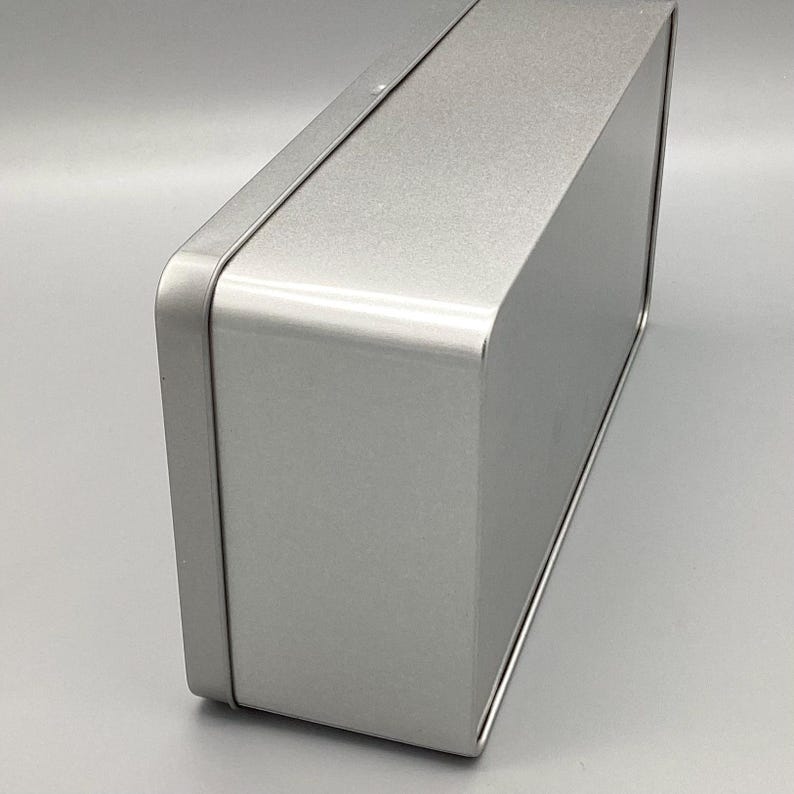 May include: A silver metal rectangular box with a smooth, shiny surface. The box is closed and has a slightly rounded edge.
