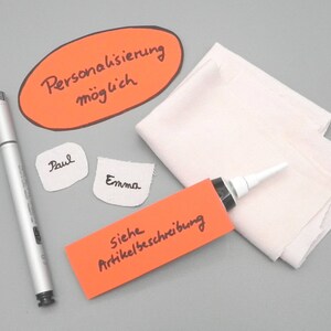 May include: An orange oval sticker with the text "Personalisierung m&ouml;glich" in black marker.  Two white fabric labels with the names "Paul" and "Emma" written in black marker. A red rectangular sticker with the text "Siehe Artikelbeschreibung" in black marker. A silver marker pen. A white fabric and a tube of glue with a white nozzle.