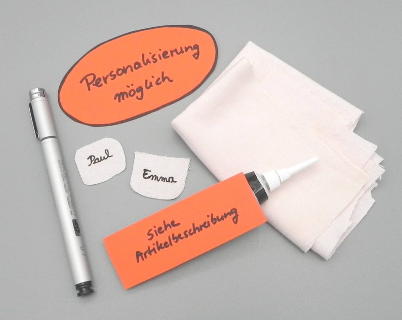 May include: An orange oval sticker with the text "Personalisierung m&ouml;glich" in black marker.  Two white fabric labels with the names "Paul" and "Emma" written in black marker. A red rectangular sticker with the text "Siehe Artikelbeschreibung" in black marker. A silver marker pen. A white fabric and a tube of glue with a white nozzle.