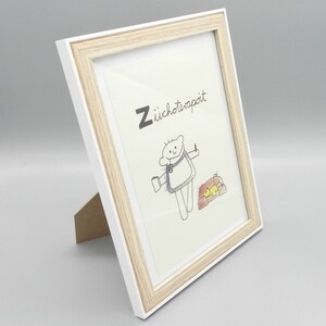 May include: A white picture frame with a light wood trim. The frame is standing upright on a white surface. The frame contains a drawing of a person with a cup and a small animal. The drawing is titled "Z&uuml;choterapit".