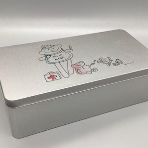 May include: A silver metal first aid kit with a cartoon illustration of a teddy bear doctor with a red cross and the text "Haus Arzt".