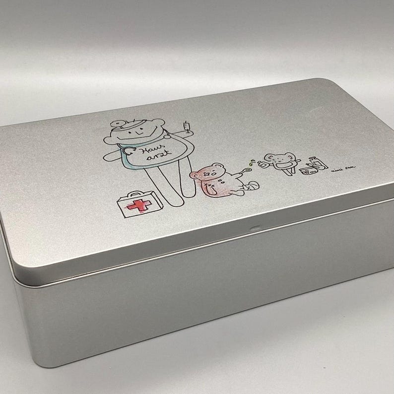 May include: A silver metal first aid kit with a cartoon illustration of a teddy bear doctor with a red cross and the text "Haus Arzt".