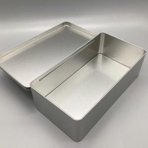 May include: A silver metal rectangular box with a hinged lid. The box is open and the inside is visible.