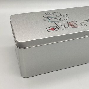May include: A silver metal tin with a cartoon drawing of a doctor with a first aid kit and a teddy bear patient. The text on the tin says "Haus Arzt".