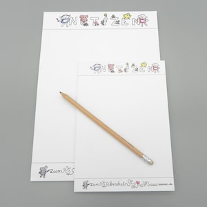 Può includere: Due blocchi note bianchi con un design di animali dei cartoni animati in bianco e nero. Il blocco note superiore ha la parola "NOTIZEN" scritta in nero con un animale dei cartoni animati sopra ogni lettera. Il blocco note inferiore ha la parola "zum" scritta in nero con un animale dei cartoni animati accanto. Una matita di legno è sopra i blocchi note.