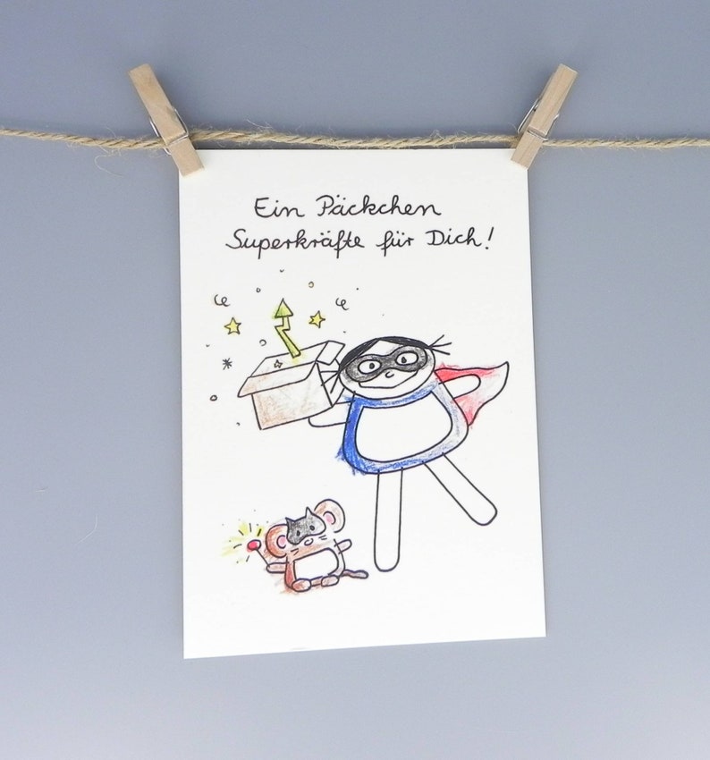 Postkarte mit nini san Mädchen in blau als Superheldin mit roten Umhang und Maske. Sie hält ein Päckchen Superkräfte (dargestellt durch einen Blitz und Sterne) in die Höhe. Eine Maus mit Maske und Zauberstab sitzt neben ihr.