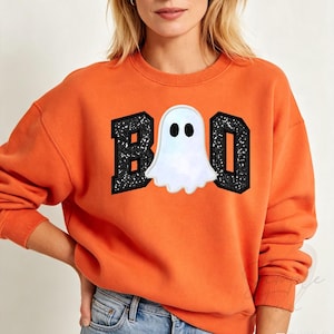 May include: An orange crewneck sweatshirt featuring a graphic design on the chest. The design displays the word BOO in large, black, glittery block letters, with a white ghost illustration replacing the letter O in the centre of the text.