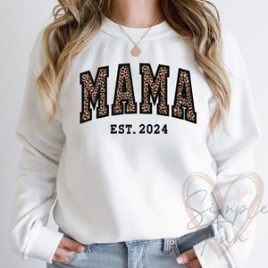 以下が含まれることがあります： 黒とヒョウ柄の「MAMA EST. 2024」のグラフィックがプリントされた白いスウェットシャツです。
