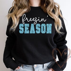 May include: Black sweatshirt with the words "freezin' SEASON" in white script and light blue block letters. The letters in "SEASON" have a glittery texture. The sweatshirt has long sleeves and a crew neck.