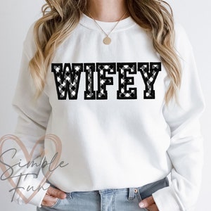 May include: White sweatshirt with the word "WIFEY" in large, bold, black and white buffalo plaid letters across the chest. The sweatshirt has long sleeves and a crew neck.
