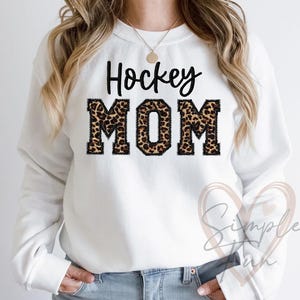 Peut inclure: Sweat-shirt blanc avec le texte "Hockey Mom" en lettres noires avec un motif léopard.