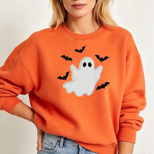 May include: An orange long-sleeve sweatshirt featuring a central graphic of a white ghost with black eyes and a mouth, surrounded by four small black flying bats arranged in a circular pattern around the ghost figure on the chest area.