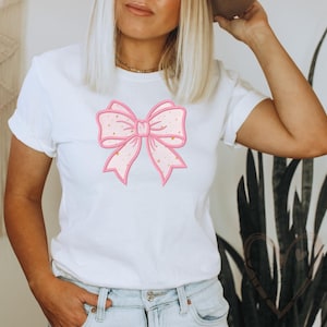 Puede incluir: Una camiseta blanca de manga corta con un gráfico grande y centrado de un lazo rosa claro con un contorno rosa más oscuro y pequeños detalles de estrellas doradas dispersos por la superficie del diseño del lazo, combinada con pantalones vaqueros azul claro.