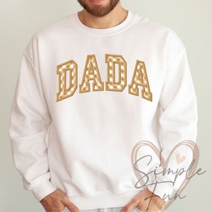Op de afbeelding: Witte trui met het woord "DADA" in bruin en wit geruite letters.