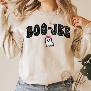 May include: Beige sweatshirt with the text "BOO-JEE" in black, outlined in white. Below the text is a white ghost with a pink bow. The sweatshirt is a casual, long-sleeved top.