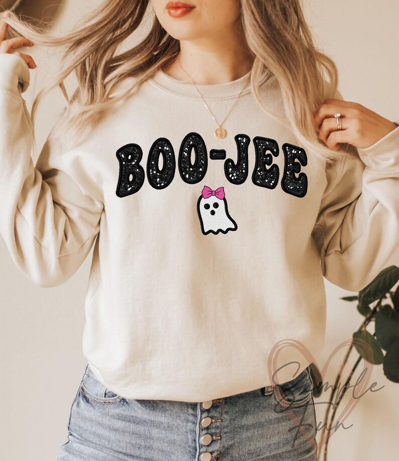May include: Beige sweatshirt with the text "BOO-JEE" in black, outlined in white. Below the text is a white ghost with a pink bow. The sweatshirt is a casual, long-sleeved top.