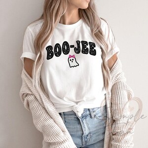 May include: White t-shirt with the text "BOO-JEE" in black, glittery letters. A small ghost with a pink bow is below the text. The shirt is paired with blue jeans and a cream-colored cardigan.