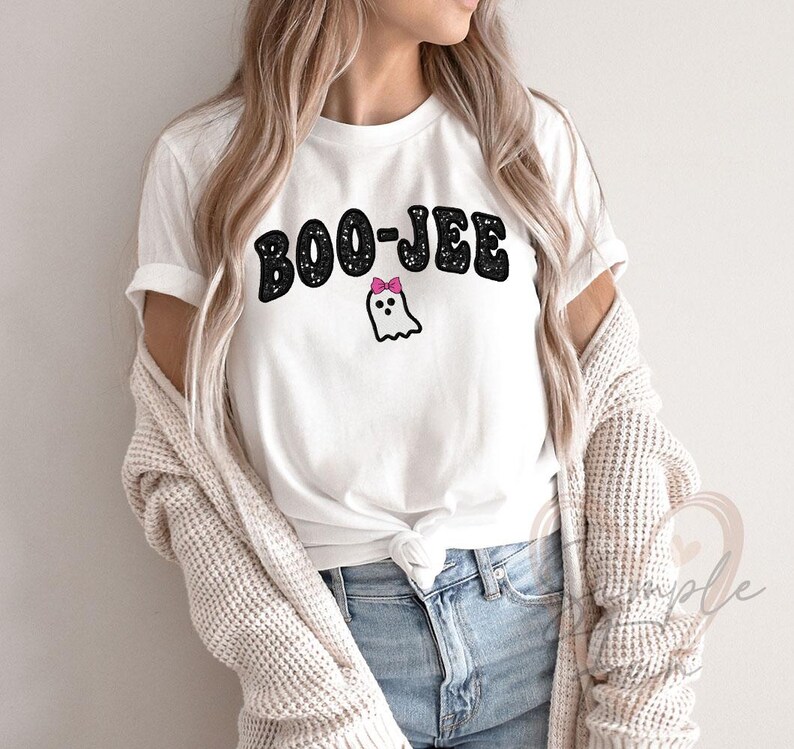May include: White t-shirt with the text "BOO-JEE" in black, glittery letters. A small ghost with a pink bow is below the text. The shirt is paired with blue jeans and a cream-colored cardigan.