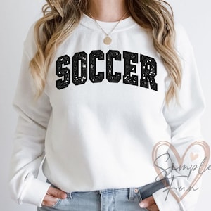 Puede incluir: Sudadera blanca con la palabra "SOCCER" en letras grandes, negras y texturizadas en el pecho. La sudadera tiene mangas largas y cuello redondo. El estilo general es casual y deportivo.