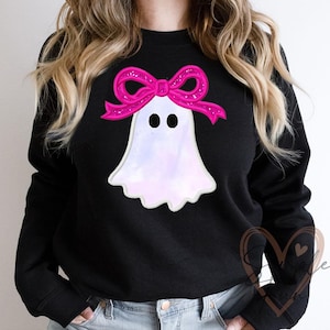 Peut inclure: Sweat-shirt noir avec un motif de fantôme et un nœud rose. Le fantôme est blanc avec un effet nacré et des yeux noirs. Le nœud est rose vif avec des paillettes. Le sweat-shirt est de style col rond.