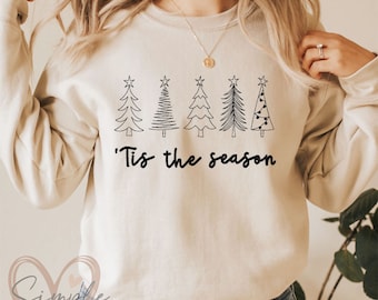 Può includere: Felpa beige con grafica nera di cinque alberi di Natale e la frase "'Tis the season". Gli alberi hanno disegni diversi e la felpa è di colore neutro.