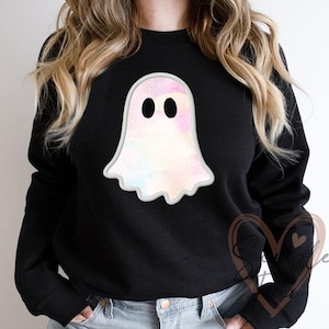 May include: Black sweatshirt featuring a watercolor ghost design. The ghost is white with pastel pink, blue, and yellow accents, and has two black oval eyes. The ghost has a white outline and a wavy bottom edge.