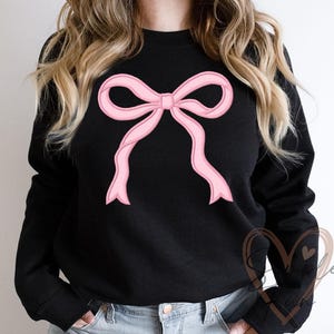 Puede incluir: Sudadera negra con un diseño de lazo rosa grande. El lazo es de color rosa claro con un contorno rosa más oscuro, creando un efecto 3D. La sudadera tiene mangas largas y cuello redondo.