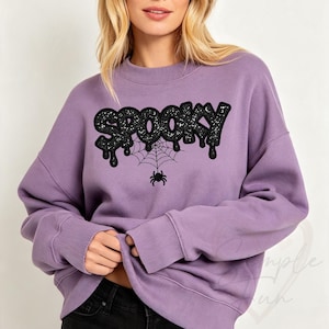 May include: A lavender crewneck sweatshirt featuring the word SPOOKY in black glittery, dripping letters. Below the text is a black spiderweb graphic with a small black spider hanging from the center, all centered on the chest of the garment.