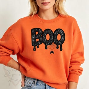 May include: An orange crewneck sweatshirt featuring the word BOO in black glittery, dripping letters. A black spider web is printed beneath the letters, with a small black spider hanging from the center of the web, centered on the chest area.