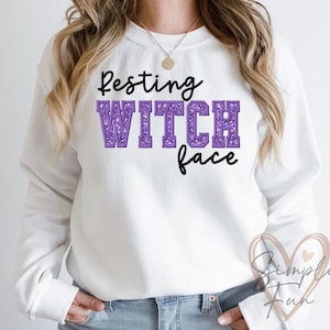 May include: White sweatshirt featuring the phrase "Resting Witch Face". The word "WITCH" is in a purple glitter font. A gold necklace is visible. The sweatshirt is a casual style, suitable for everyday wear.
