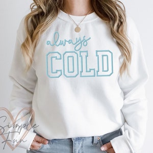 May include: White sweatshirt with the text "always COLD" in blue lettering. The text is in a varsity style font.