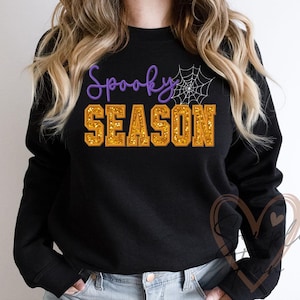 Könnte beinhalten: Schwarzes Sweatshirt mit den Worten "Spooky SEASON" in lila und goldfarbenem Glitzer. Rechts vom Wort "Spooky" befindet sich eine weiße Spinnennetz-Grafik. Das Sweatshirt hat einen Rundhalsausschnitt.