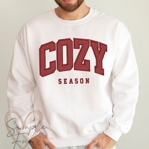 May include: White sweatshirt with the word "COZY" in maroon block letters and the word "SEASON" underneath. The sweatshirt is a crew neck style and is being worn by a person.